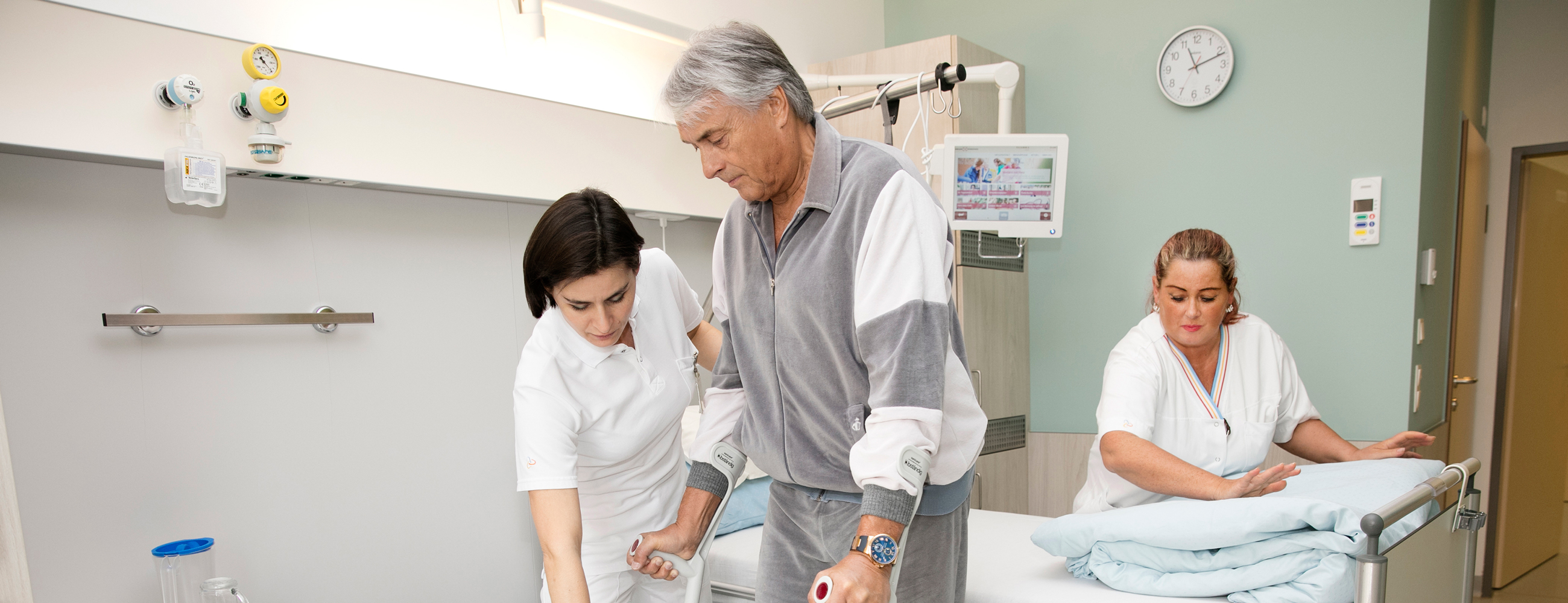 Physiotherapeutin hilft Patient mit Krücken gehen, im Hintergrund macht Pflegerin das Krankenbett