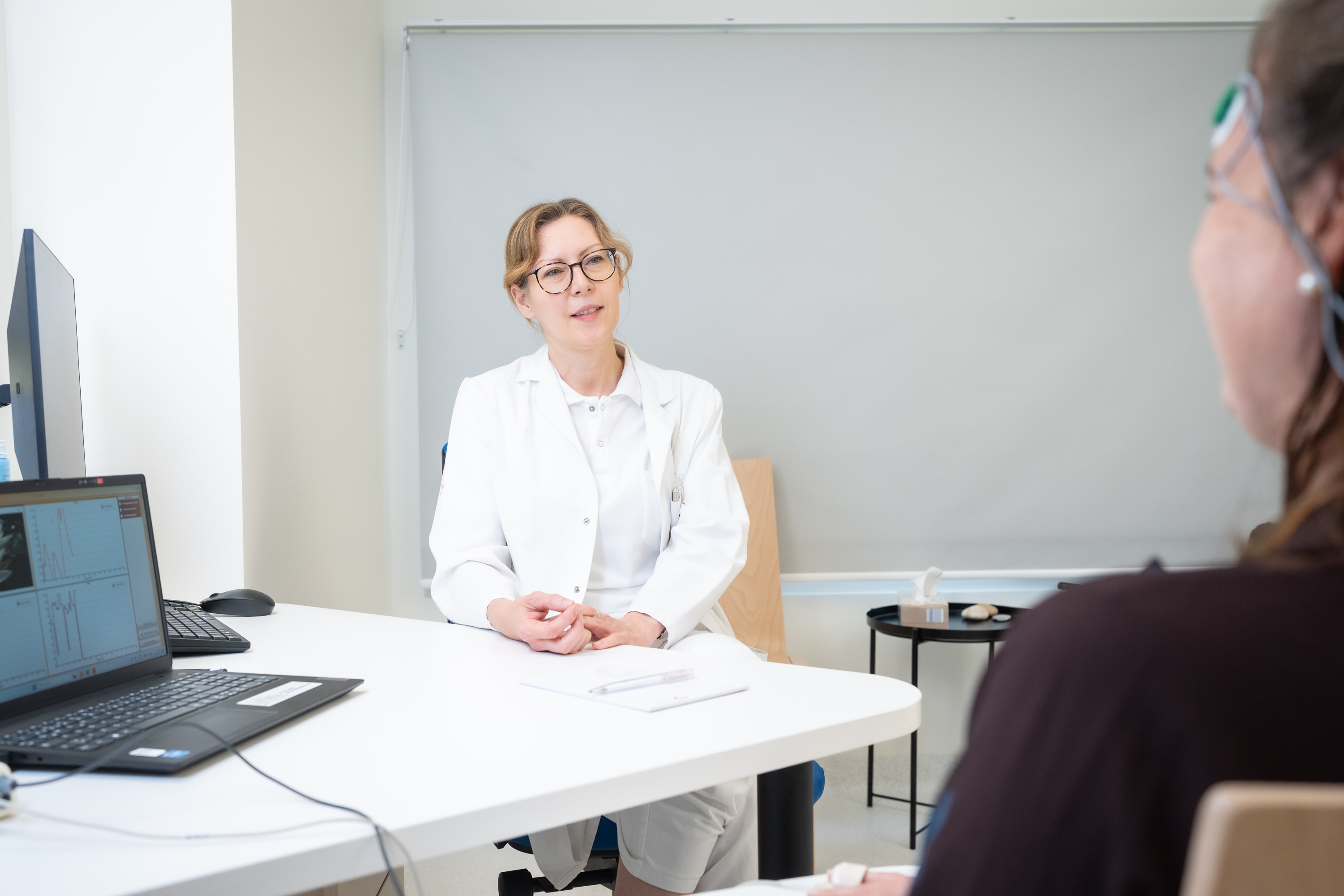 Eine Psychologin in weißer Kleidung sitzt an einem Tisch und führt ein Therapiegespräch mit einer Patientin.